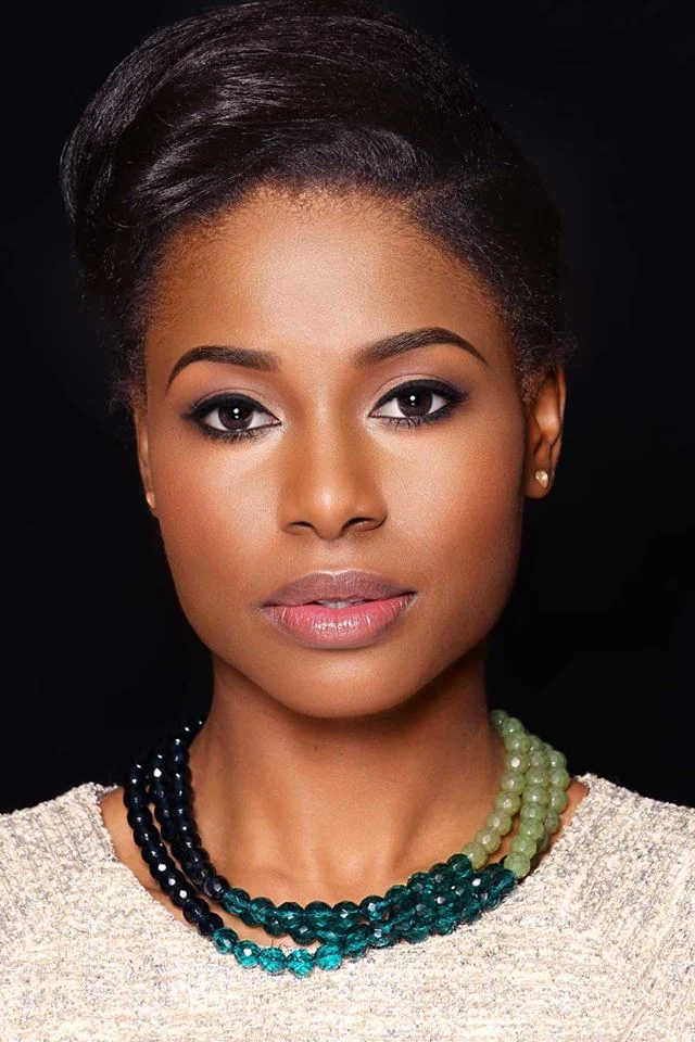 A portrait of a young African American woman with styled dark hair, wearing pearl earrings, a multi-strand necklace with various shades of green and blue beads, and a light-colored top against a black background.