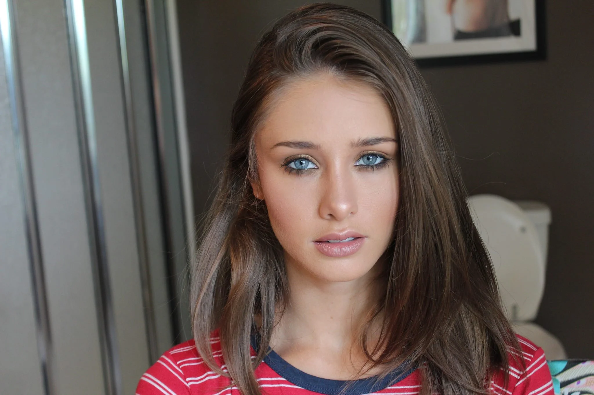 A young woman with blue eyes and long brown hair wearing a red striped shirt with a navy blue neckline, standing indoors near a mirror or glass panel.