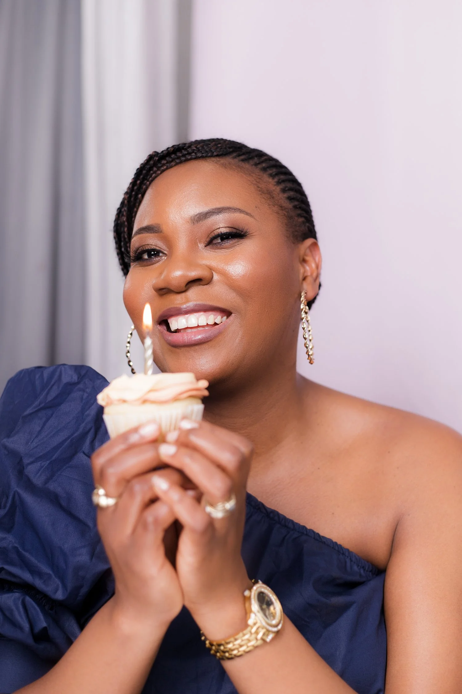 A woman with braided hair, wearing earrings and a watch, holding a cupcake with a lit candle, smiling, with a blurred background.