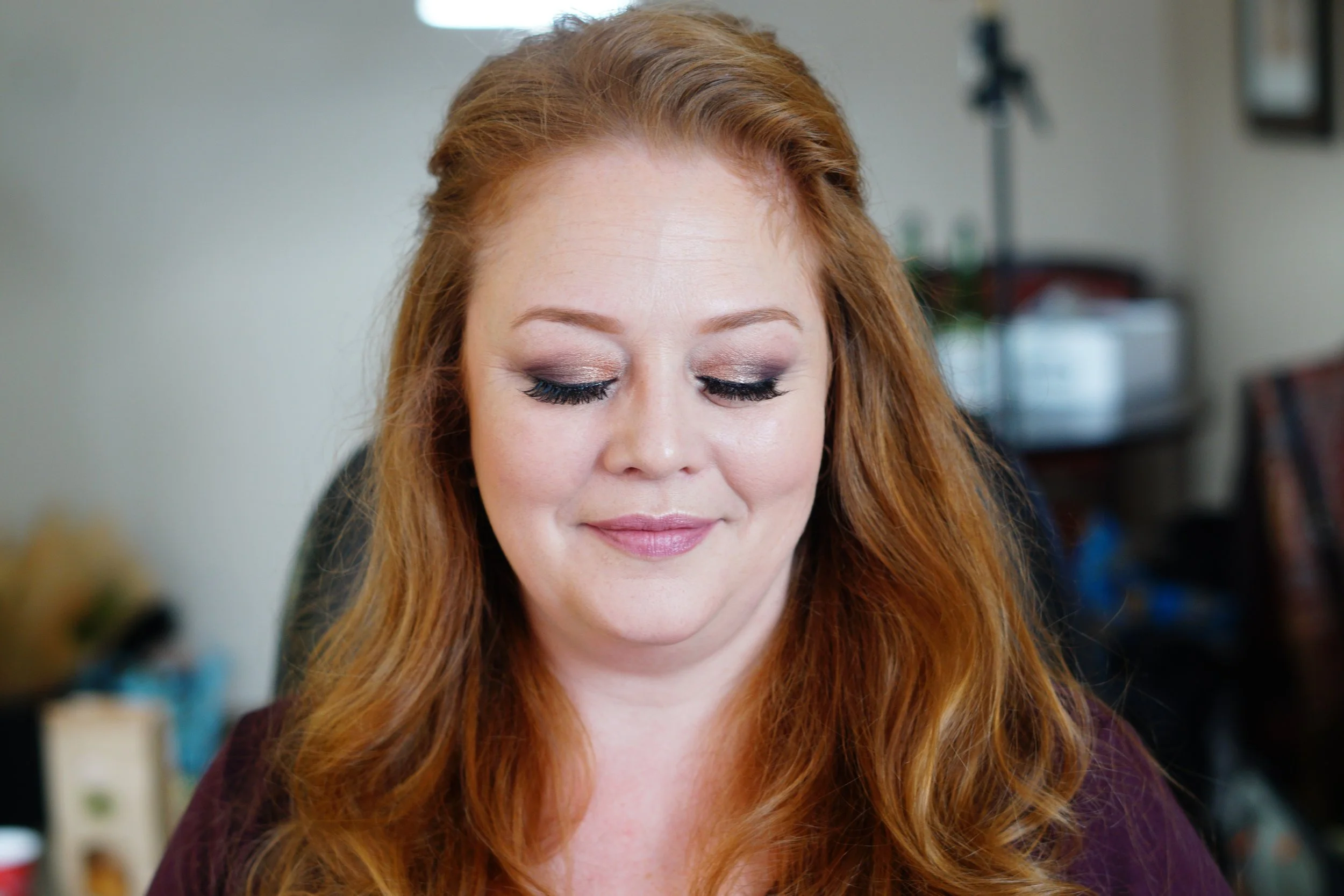 A woman with long red hair and closed eyes, smiling softly. She has makeup on with eyeshadow and eyelash extensions. The background shows a blurry room with shelves and some objects.