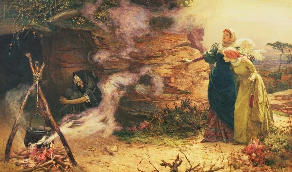 Two women are standing outside near a rocky formation, with one woman pointing towards a man inside a cave. The man is sitting near a fire with a pot hanging above it.
