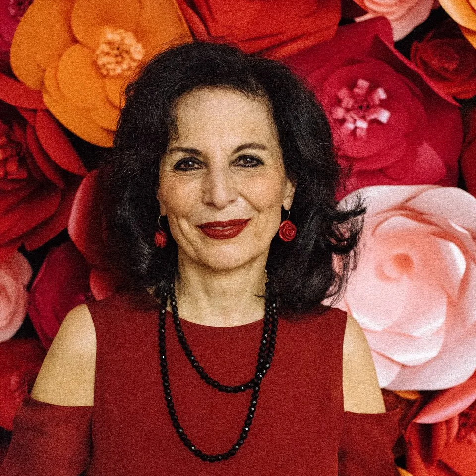 Photograph of Louise Fili, smiling in front of a backdrop of oversized paper flowers in shades of red, orange, and pink