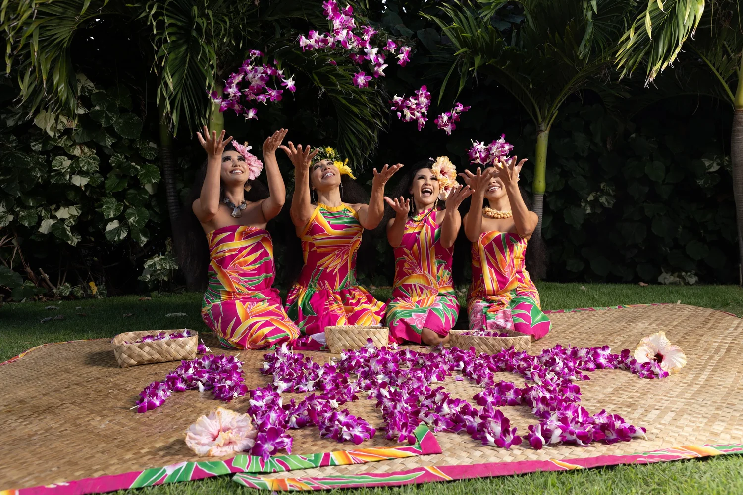 Four Malu Productions performers in vibrant Polynesian costumes tossing purple flowers upward