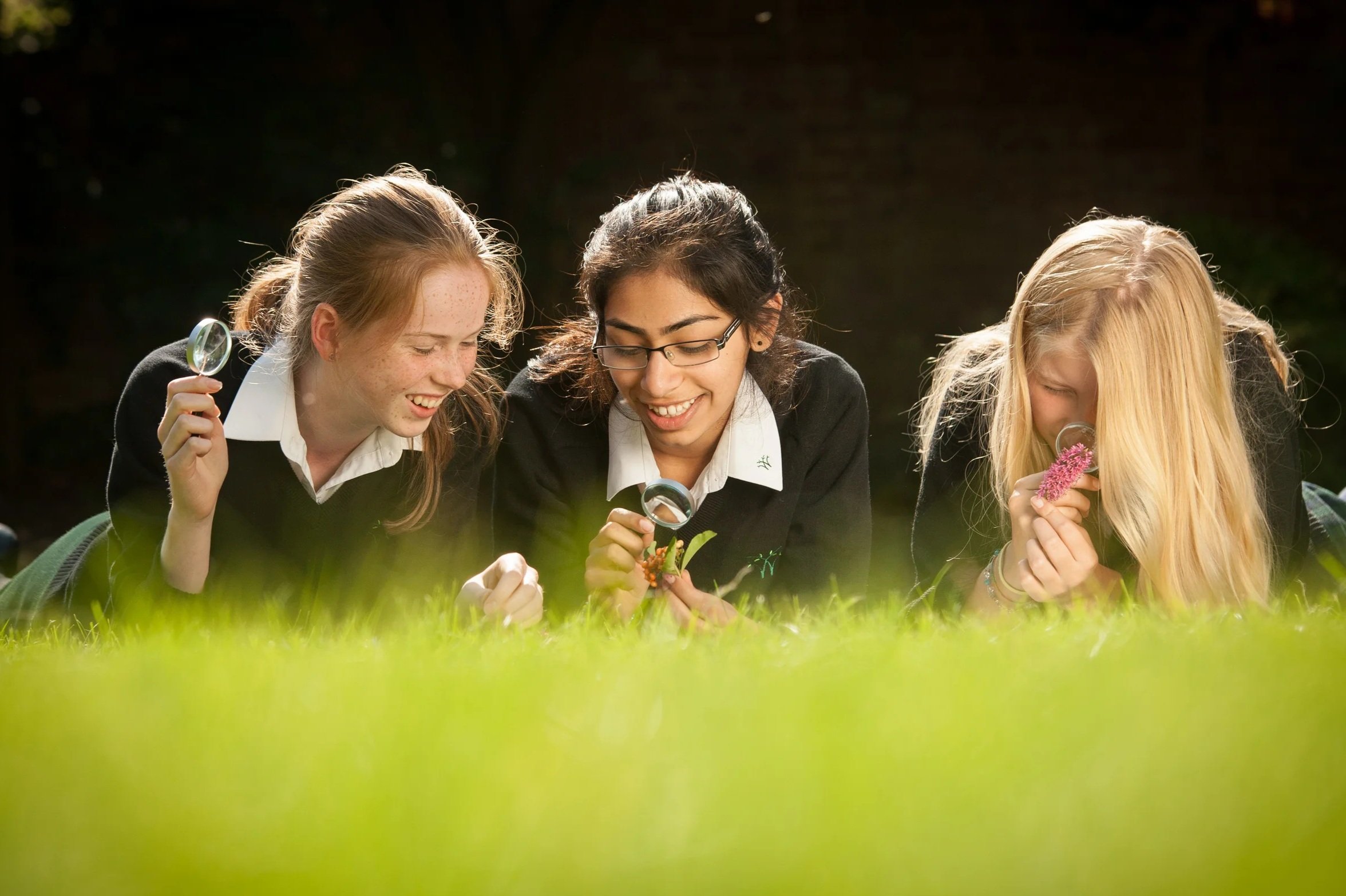 A bright lifestyle photograph for a senior school prospectus, capturing three teenage pupils in uniform sitting and laughing together on the grass during break time on a spacious school campus.
