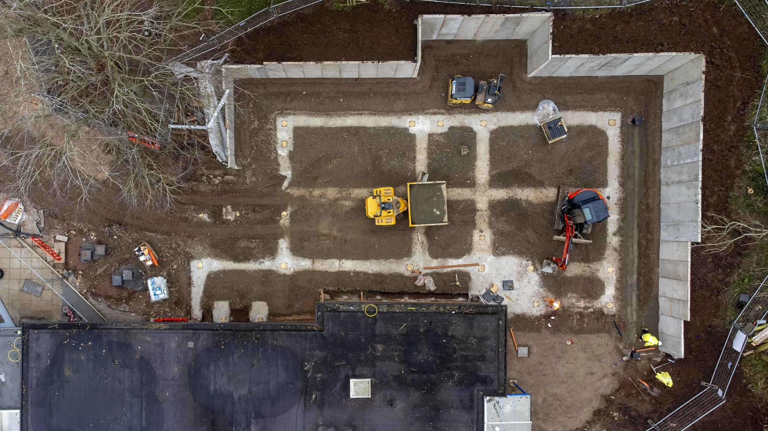 A direct top-down drone photograph of an active building site, capturing the clear layout of the construction zone, site access, and materials with architectural precision.