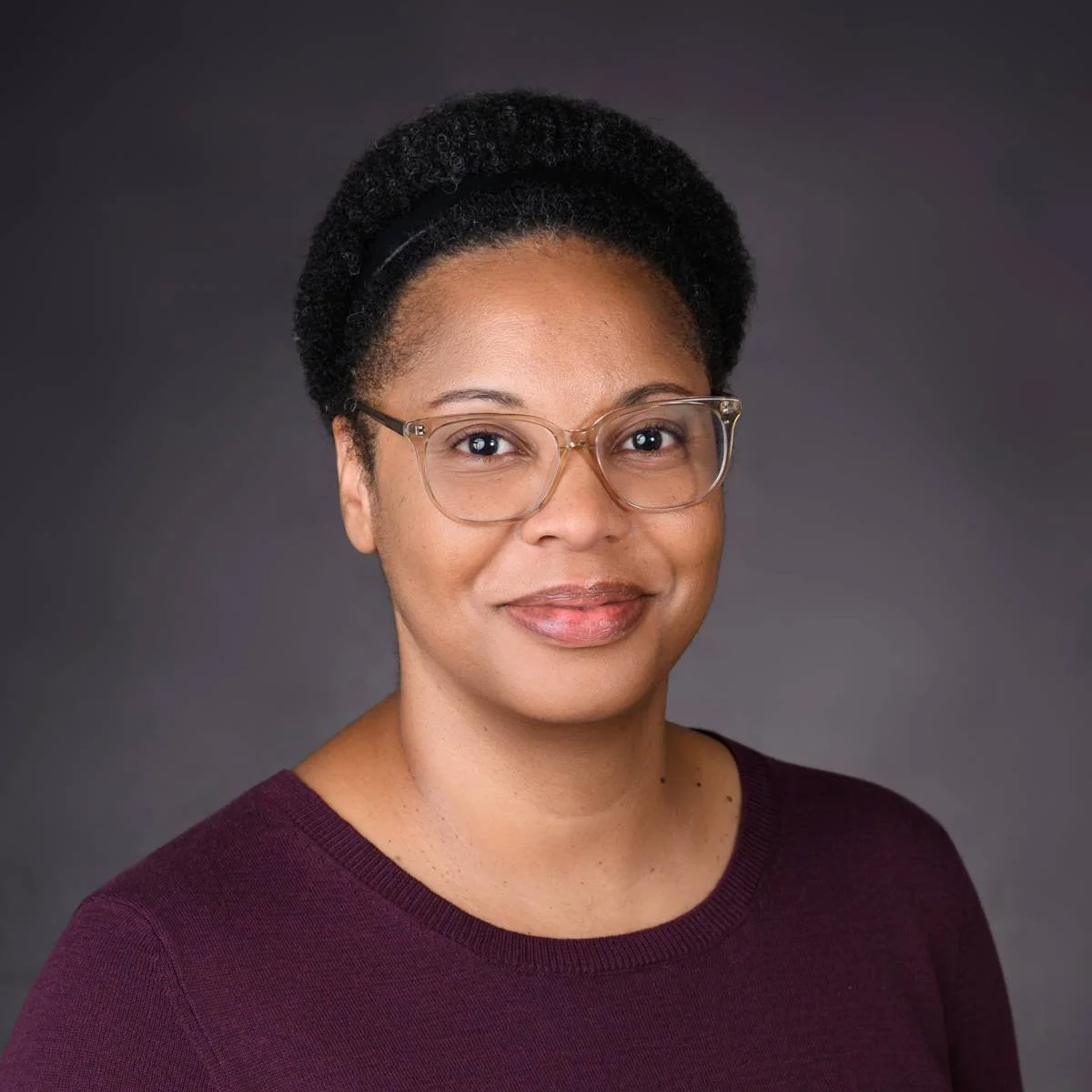 A woman with glasses and a natural hairstyle, wearing a maroon top, smiling against a dark gray background.