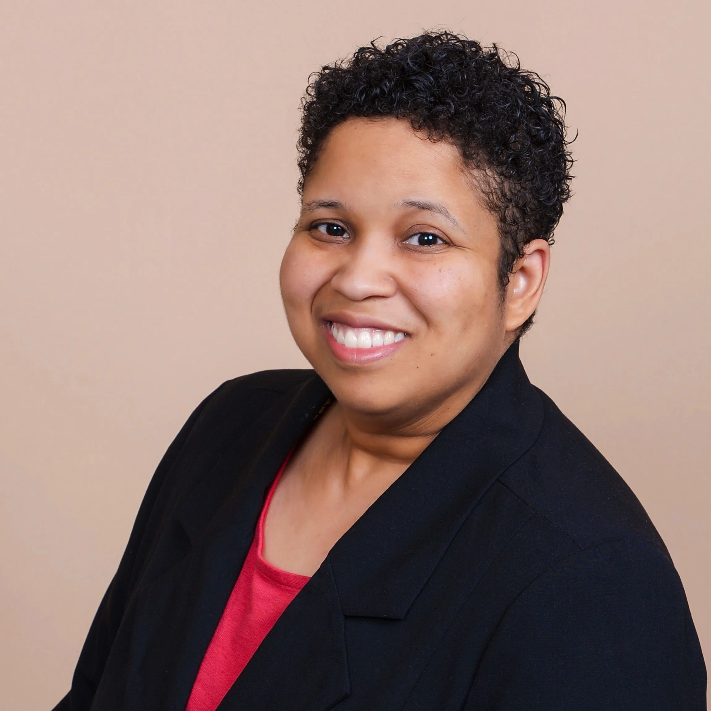A woman with short curly black hair, smiling, wearing a black blazer over a pink top, against a neutral beige background.