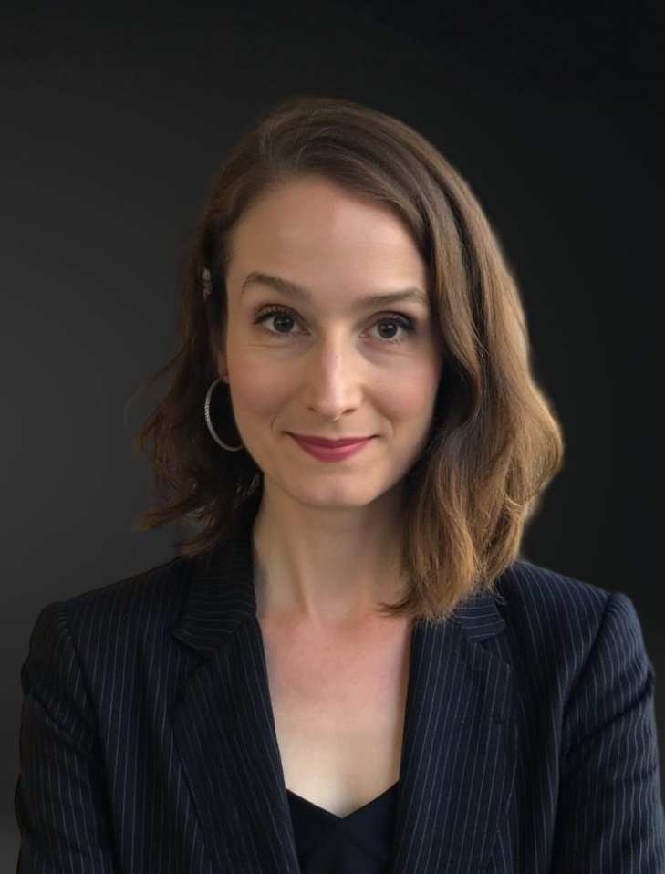 A woman with shoulder-length brown hair, wearing a pinstripe blazer, hoop earrings, and makeup, smiling at the camera against a dark background.