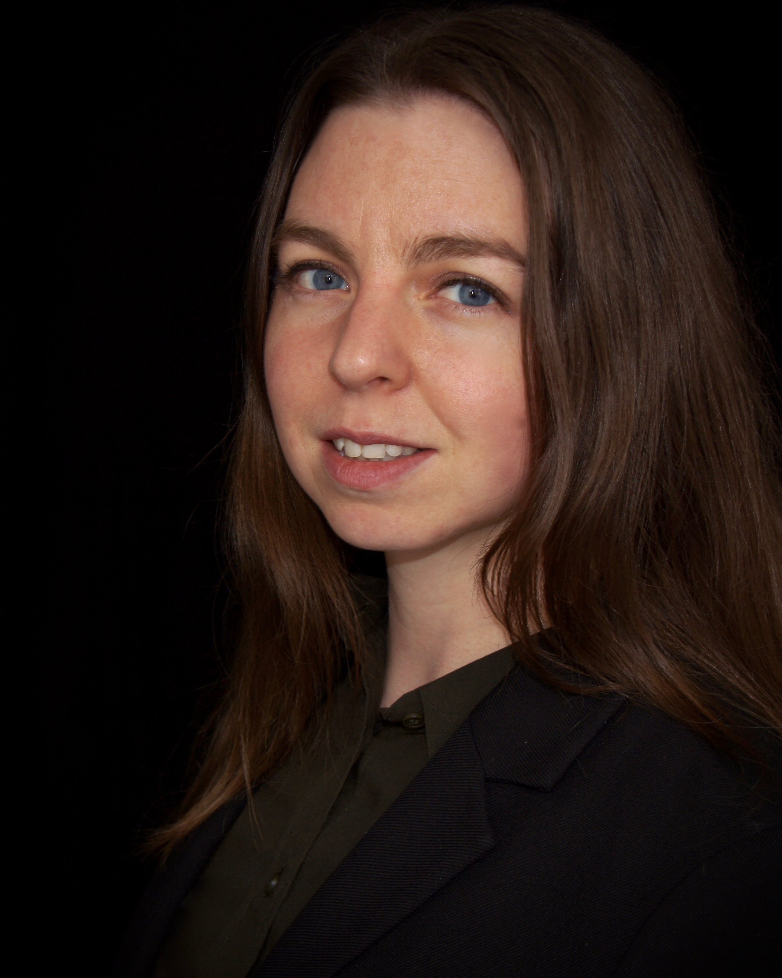 Close-up of a young woman with long brown hair and blue eyes, wearing a black blazer, against a black background.