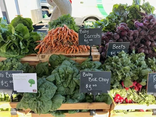 Fresh vegetables including baby carrots, beets, baby bok choy, and broccoli on display at a market stall, each with price signs.