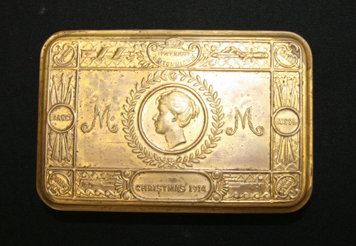 The Princess Mary tin is a flat rectangular gold coloured tin, stamped on the lid with profile of a young woman's head at the centre, letters M at left and right. Border of the lid is stamped with 'Imperium Britannicum', 'Christmas 1914' then the nations, Belgium, Japan, Russia, Monte Negro, Servia, and France.