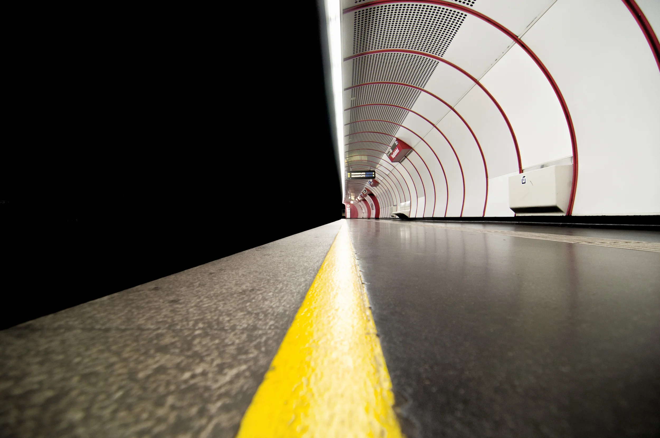Blick in eine U-Bahn-Haltestelle mit gelber Linie auf dem Boden, weißen Wänden mit roten Bögen und einer Anzeigetafel in der Mitte.