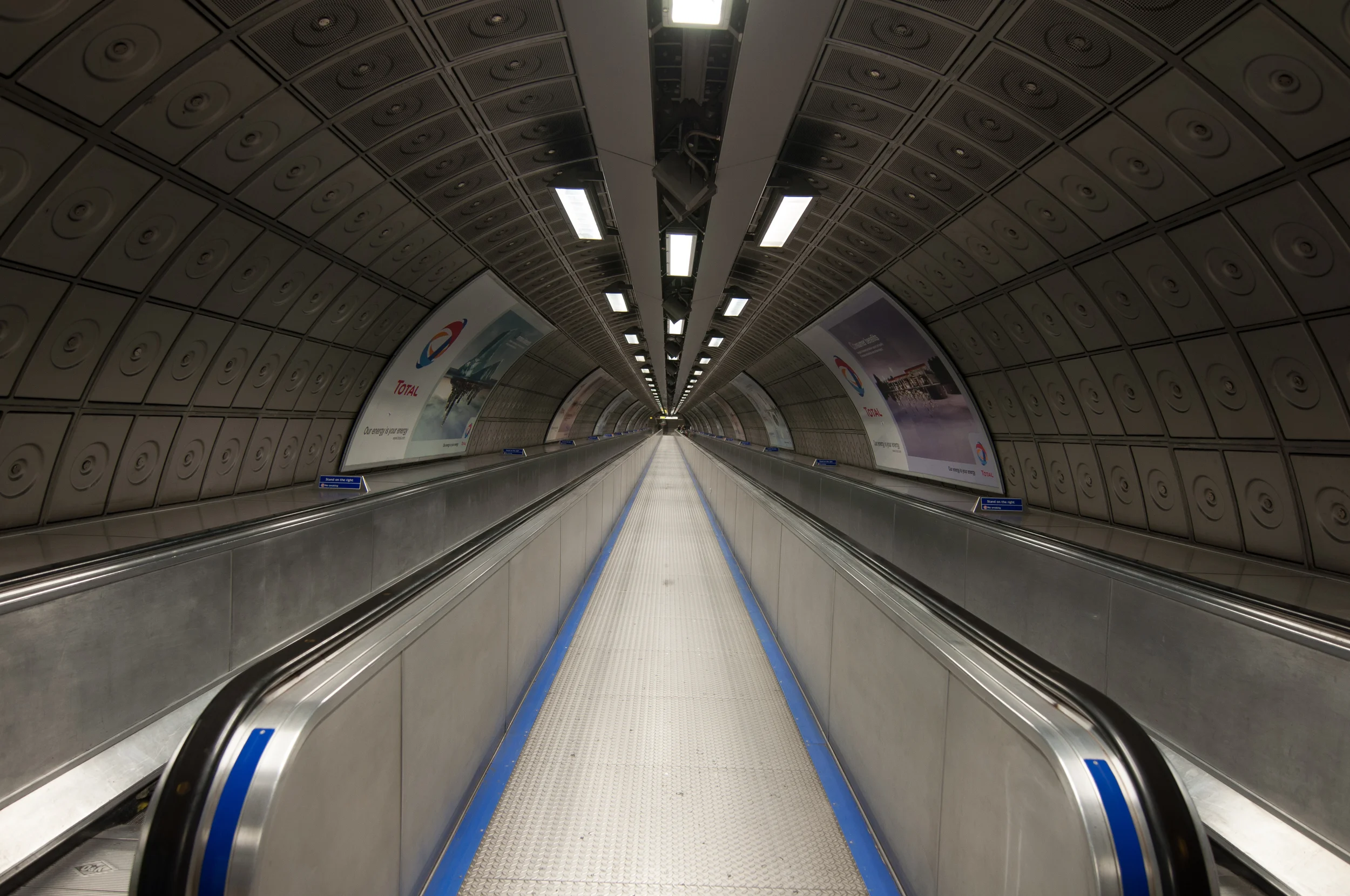 Leere Rolltreppe in einer U-Bahn-Station, Tunnel mit blauen Warnlinien am Boden und Werbeplakate an den Wänden.