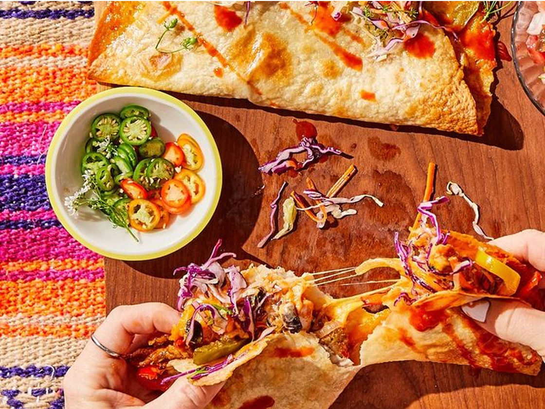 Cheese-filled taco being pulled apart, topped with slaw and hot sauce, with sliced jalapeños on the side.