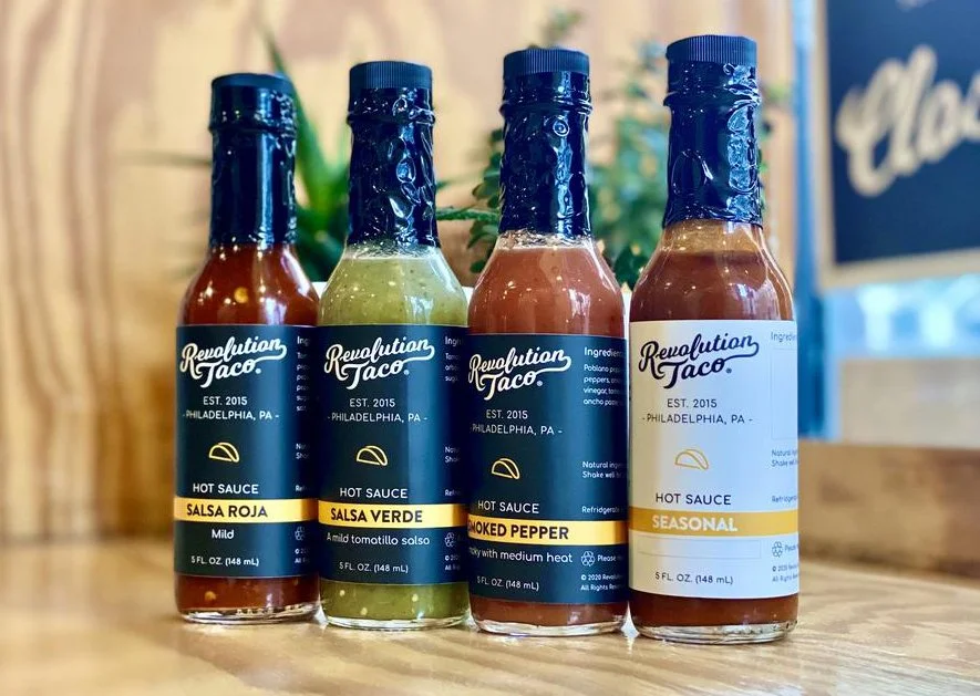 Four bottles of Revolution Taco hot sauce on a wooden counter, including salsa roja, salsa verde, smoked pepper, and seasonal.