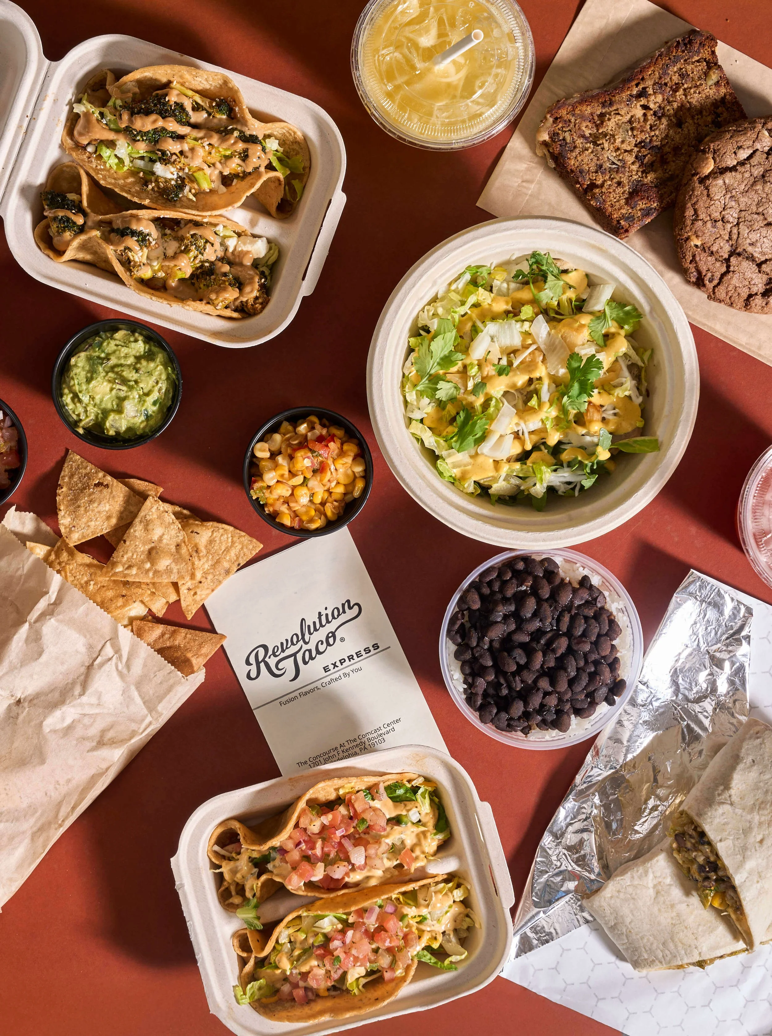 Assorted Revolution Taco Express meal spread with tacos, sides, chips, guacamole, and a burrito on a red table.