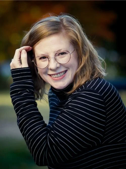 Kayce Hodos, a perinatal mental health specialist in Wake Forest NC, smiling in a headshot outdoors with soft greenery in the background, wearing round glasses and a black striped top while gently touching her hair.