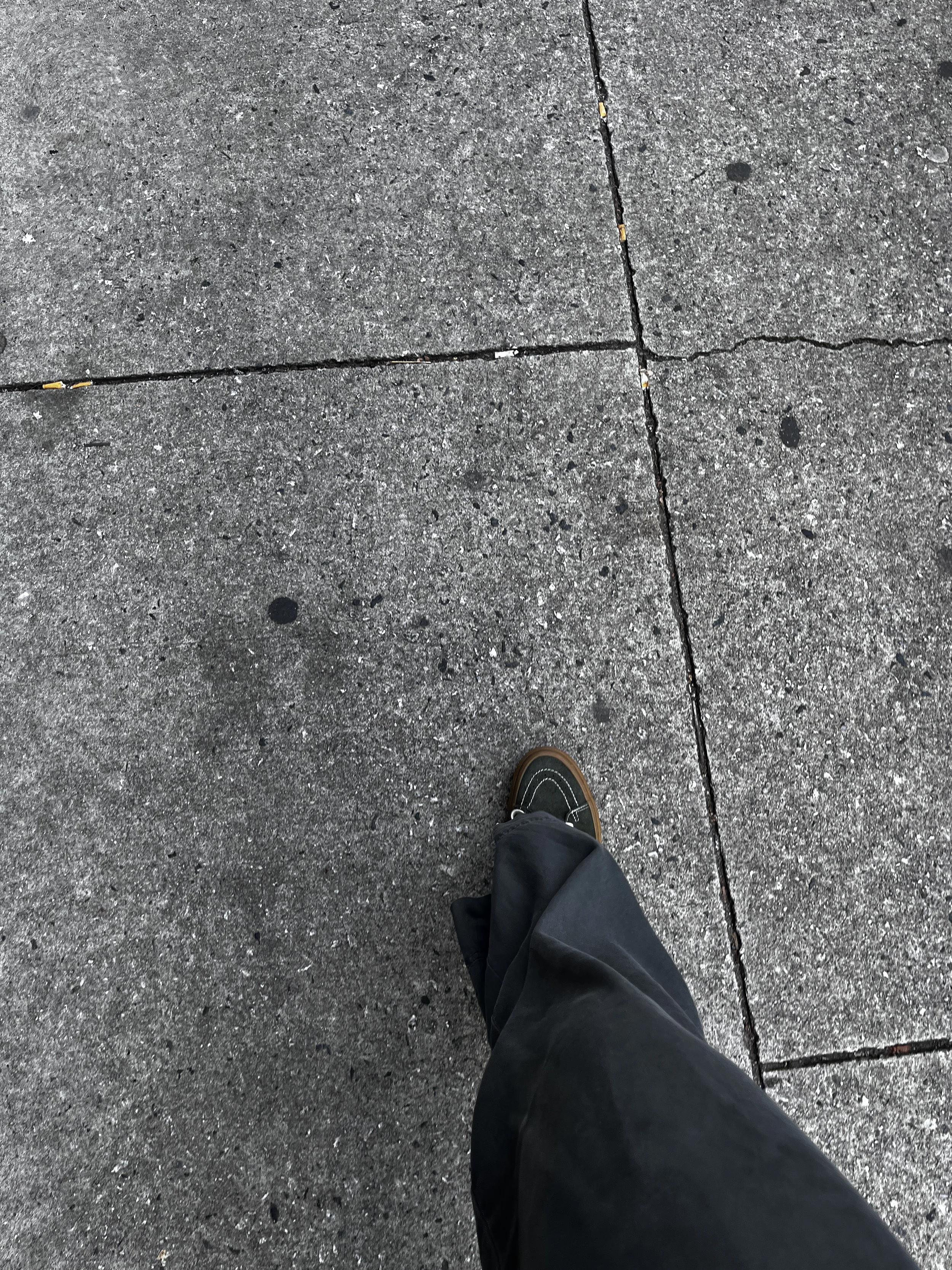 Top-down view of a person's foot walking on a concrete sidewalk with visible cracks, wearing black shoes and black pants.