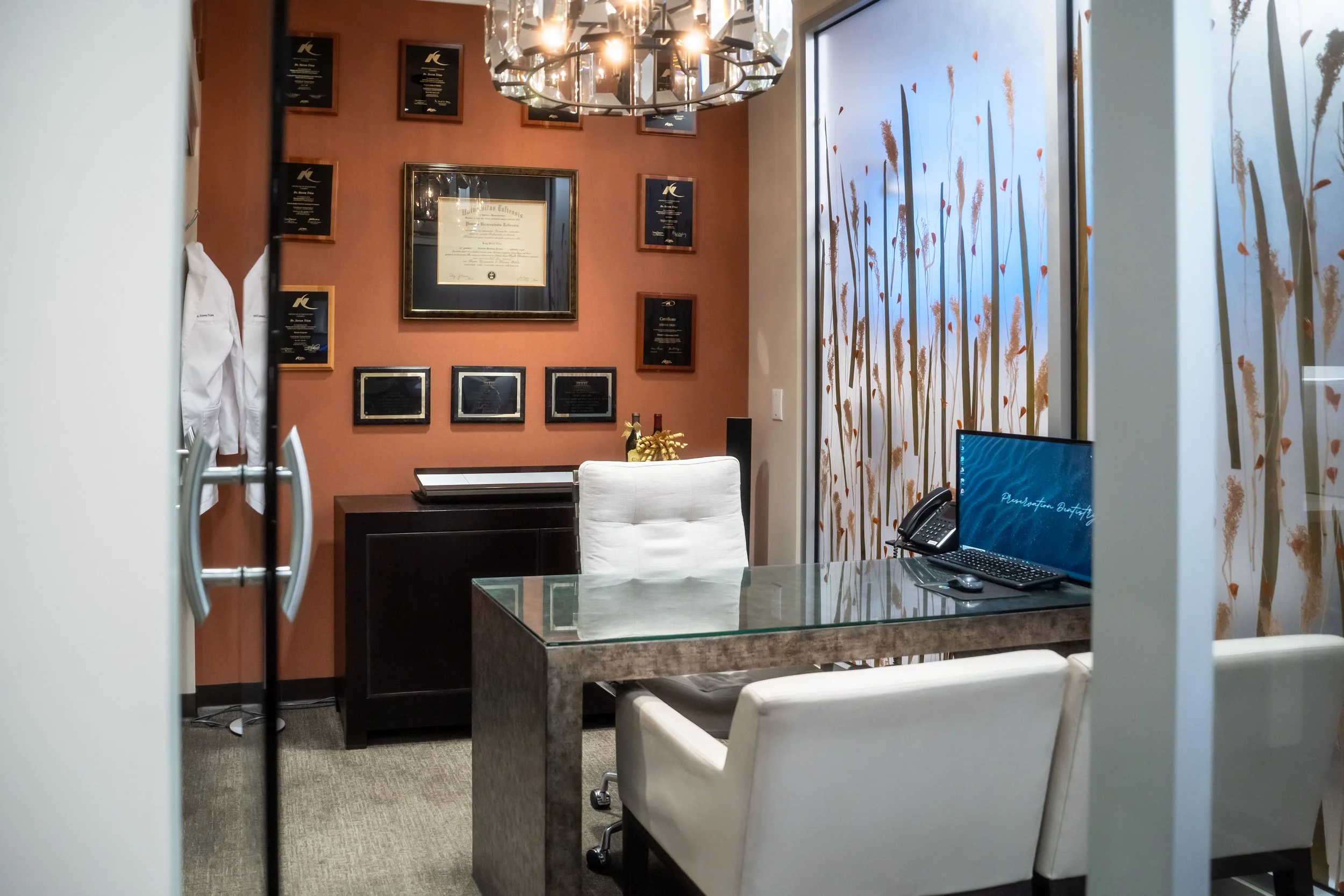 An office with framed awards and certificates on the wall, a glass desk with a computer monitor, a white office chair, and decorative window panels with a plant motif.