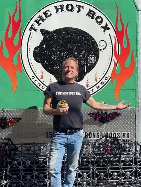 Man standing in front of a food truck called The Hot Box, holding a burger and smiling. The truck has a large logo with flames and a black bomb, with the text 'The Hot Box' and 'Genuine Fire 'N' Eat BBQ'.