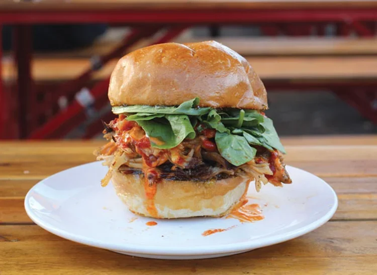 Close-up of a gourmet burger with lettuce, shredded meat, and sauce on a white plate, on a wooden table.