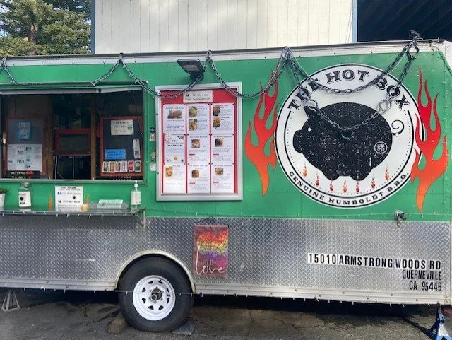 Green food truck with a black pig roasting logo, menu posted on the side, and flames graphic. Located at 15010 Armstrong Woods Rd, Guerneville, CA 95446.