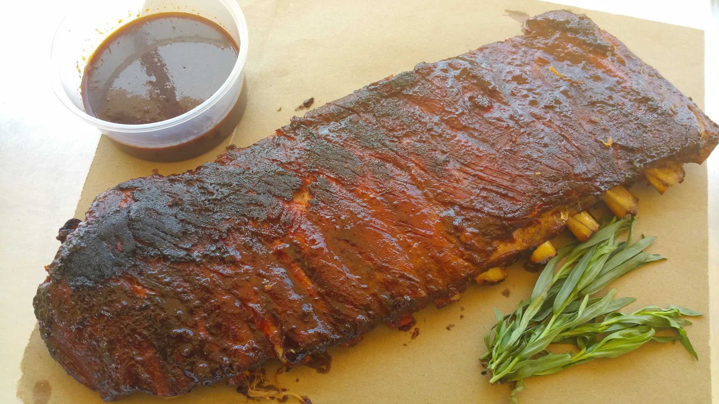 Barbecued ribs with a side of green herbs, accompanied by barbecue sauce in a small container.