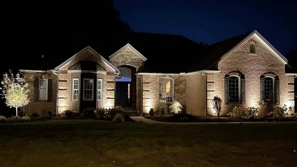 Illuminated brick house at night with landscaped yard, trees, and dark sky in the background.