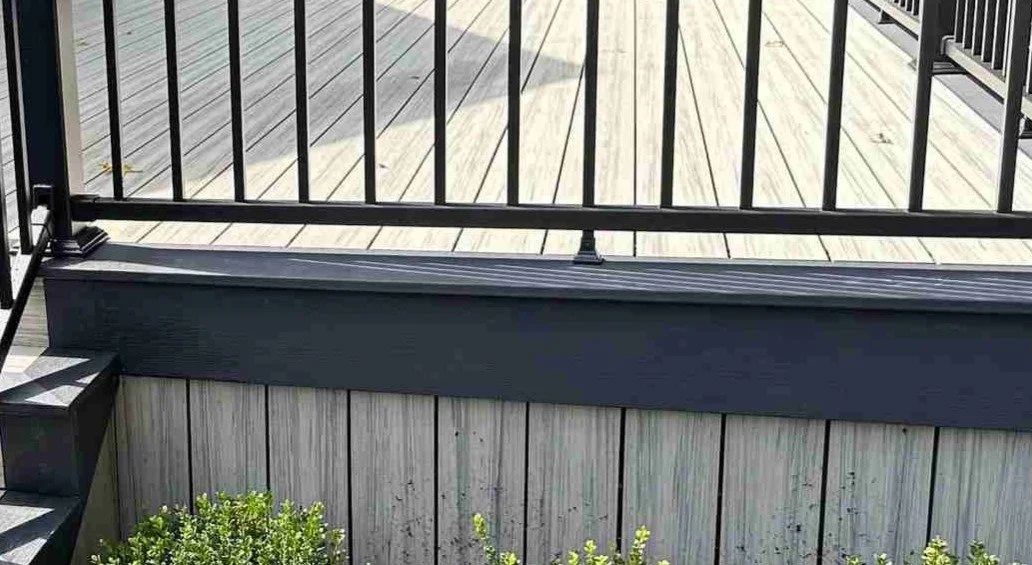 View of a composite deck with black metal railing and stairs, surrounded by a garden with green plants.