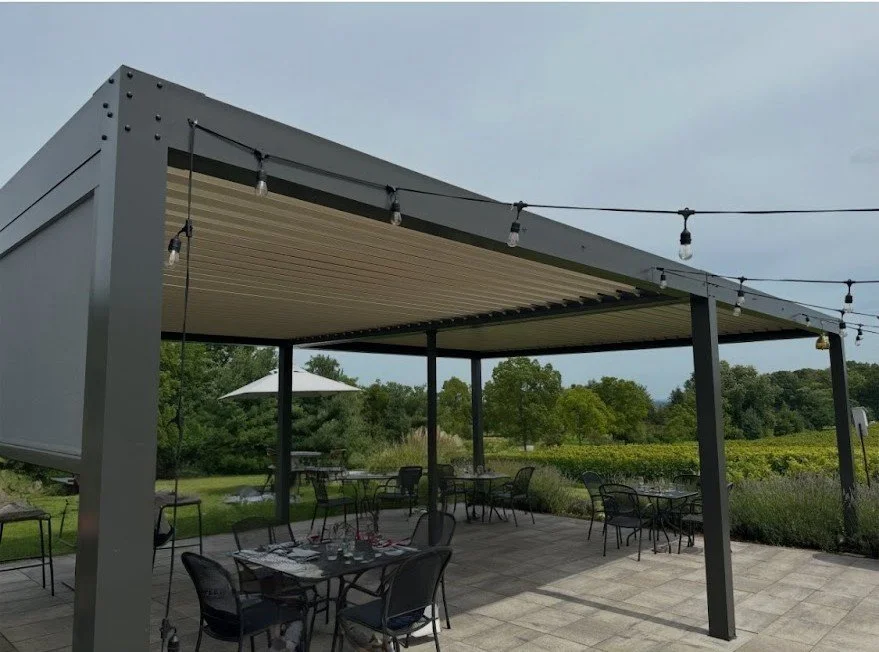 Outdoor patio with a modern gray pergola, string lights, and black metal tables and chairs surrounded by greenery and a partly cloudy sky.