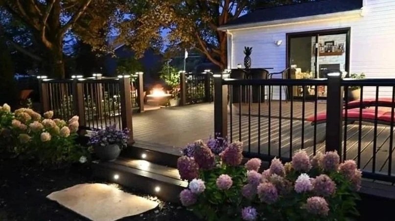 A cozy backyard deck at dusk with purple hydrangeas in front, a small fire pit glowing in the background, and a white house with a sliding door. Decorated with cushions and string lights.