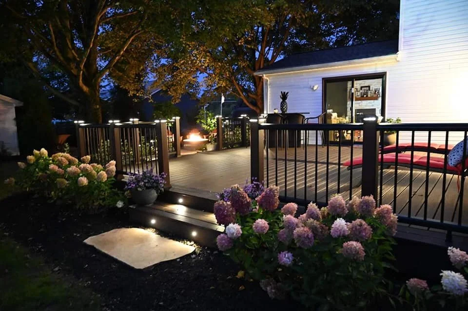 A backyard deck at dusk with string lights, potted plants, flowers, and a fire pit in the background, surrounded by trees and a white house with sliding glass door.