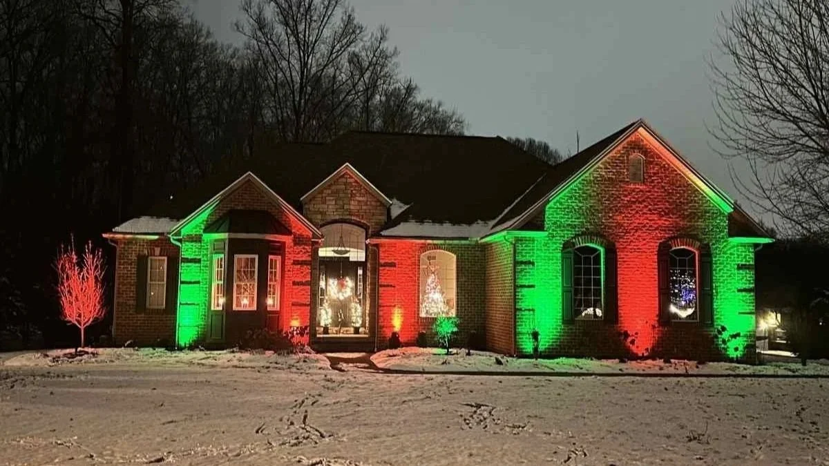 A house with Christmas lights in red and green, surrounded by snow on the ground, trees, and a cloudy sky at night.
