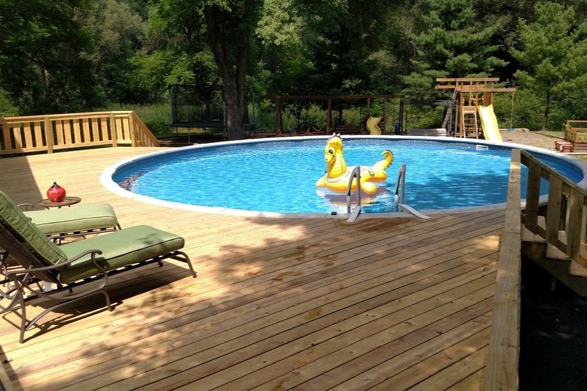 An outdoor swimming pool with a wooden deck area surrounded by trees. The pool has a yellow rubber duck float, a ladder, and a small play structure with slides in the background. Two green lounge chairs are on the deck with a small red decorative ite