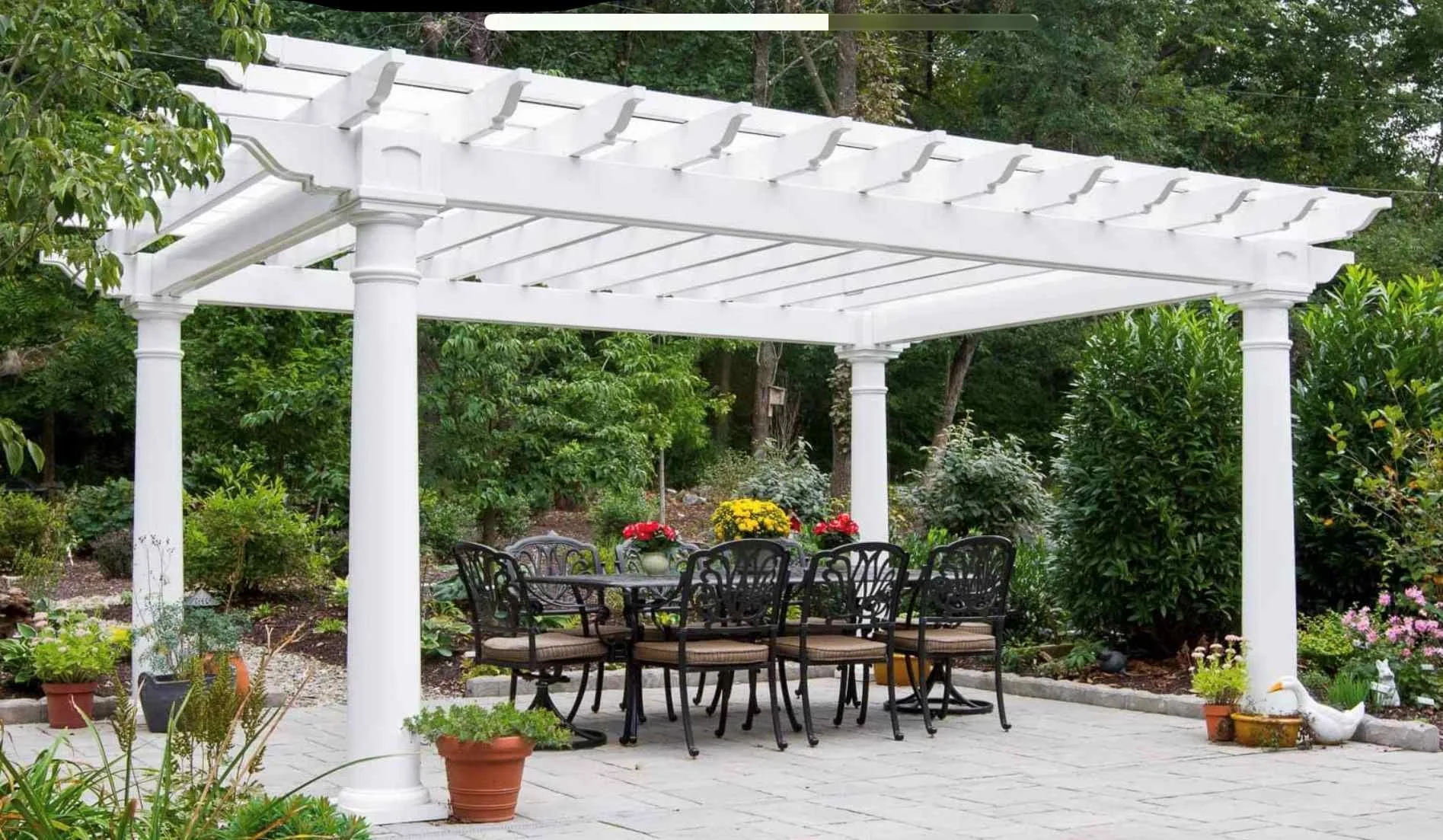 An outdoor patio with a white pergola, a black dining table with six matching chairs, surrounded by potted plants and garden shrubs, with trees in the background.