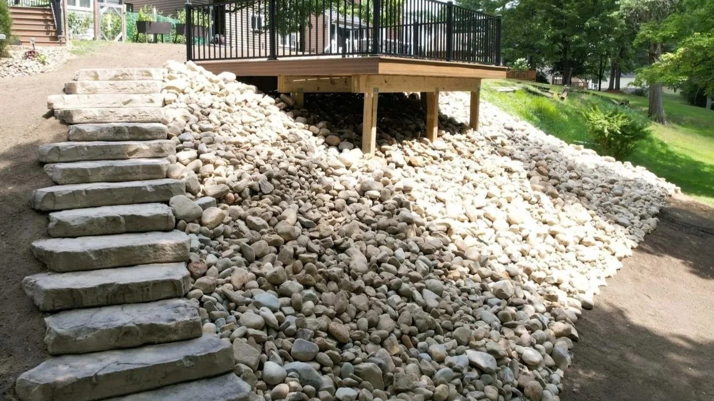 A backyard with a raised wooden deck supported by wooden posts, with Lampus stepping stone steps and a gravel slope underneath, surrounded by green grass and trees.
