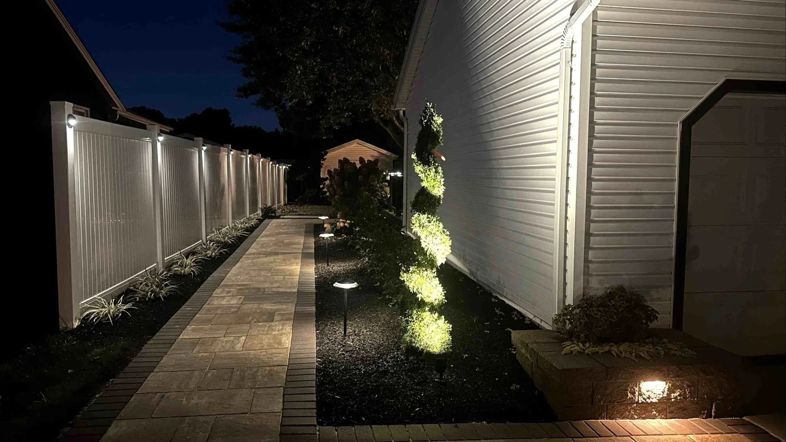 Nighttime view of a backyard with a stone pathway, small plants, and outdoor lighting next to a white house with siding. A tall white fence runs along the left side of the yard.