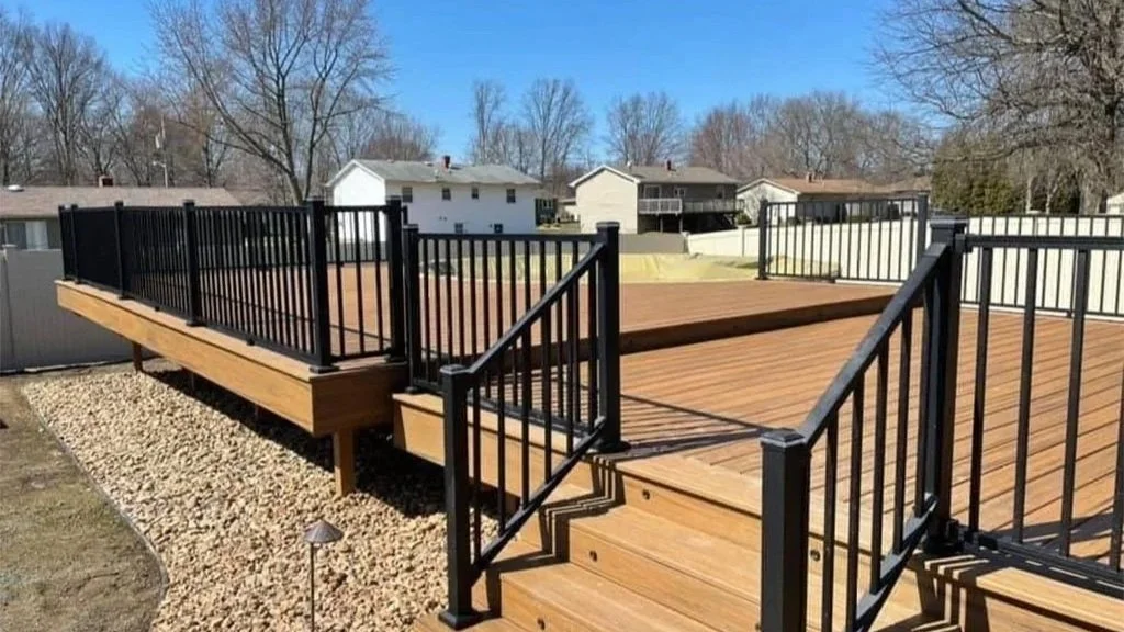 A composit deck with black metal railings, stairs leading up to the deck, and a gravel area underneath. Residential houses and leafless trees are visible in the background under a clear blue sky.