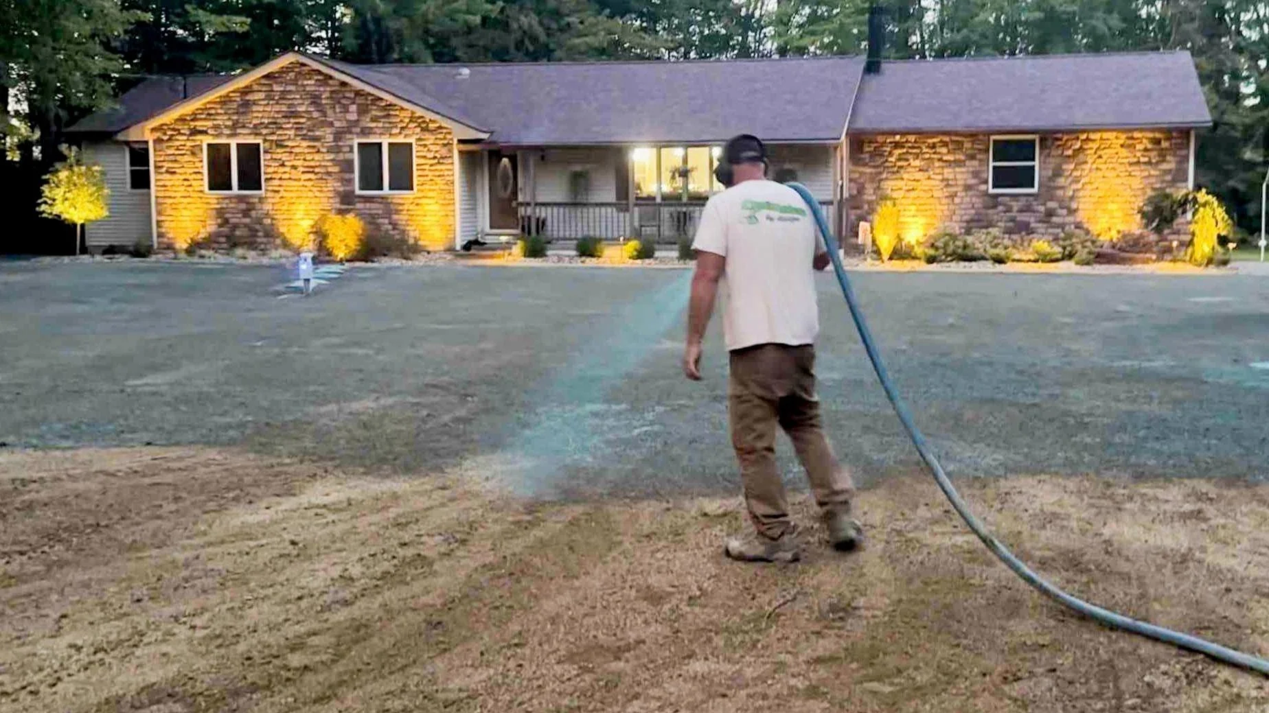 A man watering a newly seeded yard in front of a house with exterior lights on.