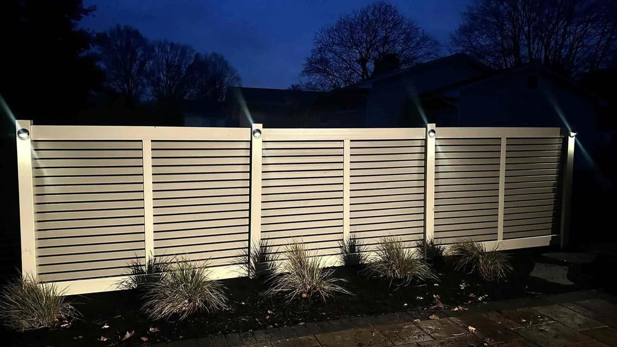 Nighttime view of a modern horizontal slat fence with four lights shining down, small plants in front, and trees and houses in the background.