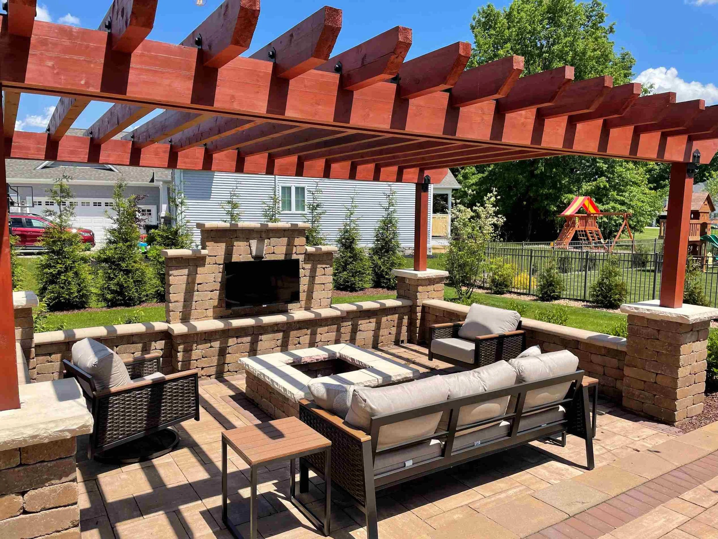 Outdoor patio with brick fireplace, seating area with cushions, under a wooden pergola, with green trees and a playground in the background.