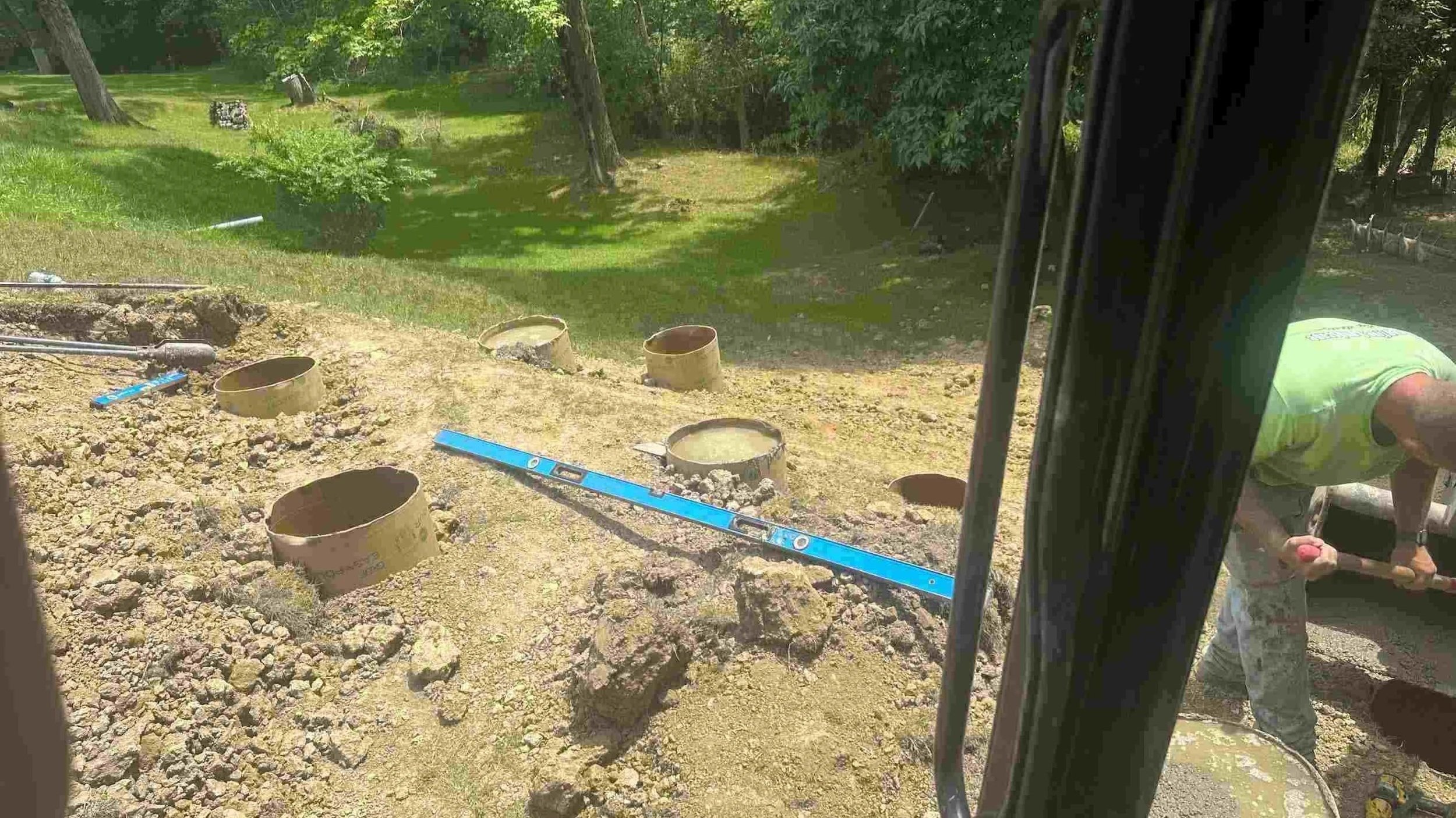 Construction worker pouring concrete at an outdoor construction site, with sewer pipes installed and level tools on ground, in a green grassy area with trees.