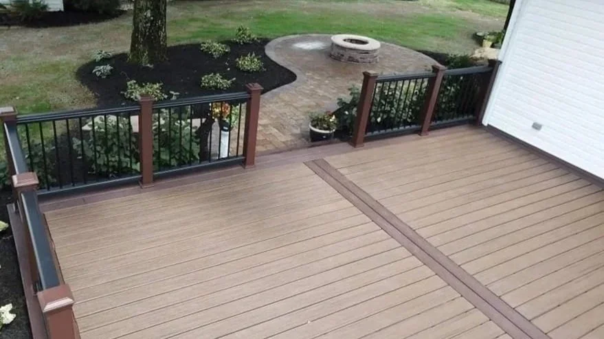 A spacious composite deck with a black and brown railing, overlooking a landscaped backyard with flowers, a tree, a stone fire pit, and a brick pathway.