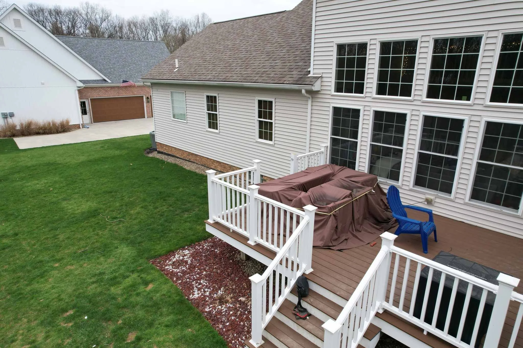 Backyard deck with a brown covered grill, a blue chair, and a white railing. Green grass surrounds the deck, and there are multiple windows on the house's exterior wall.