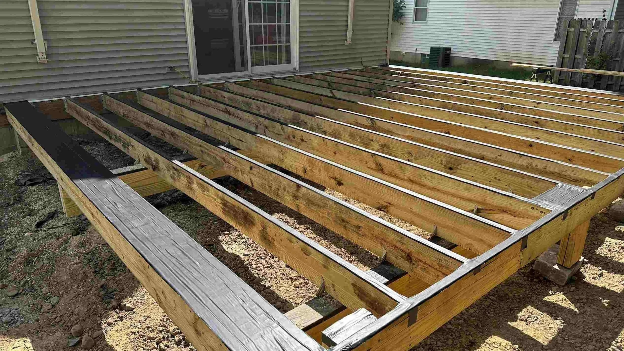 Wooden deck frame under construction in backyard, with a house and a fence in the background.