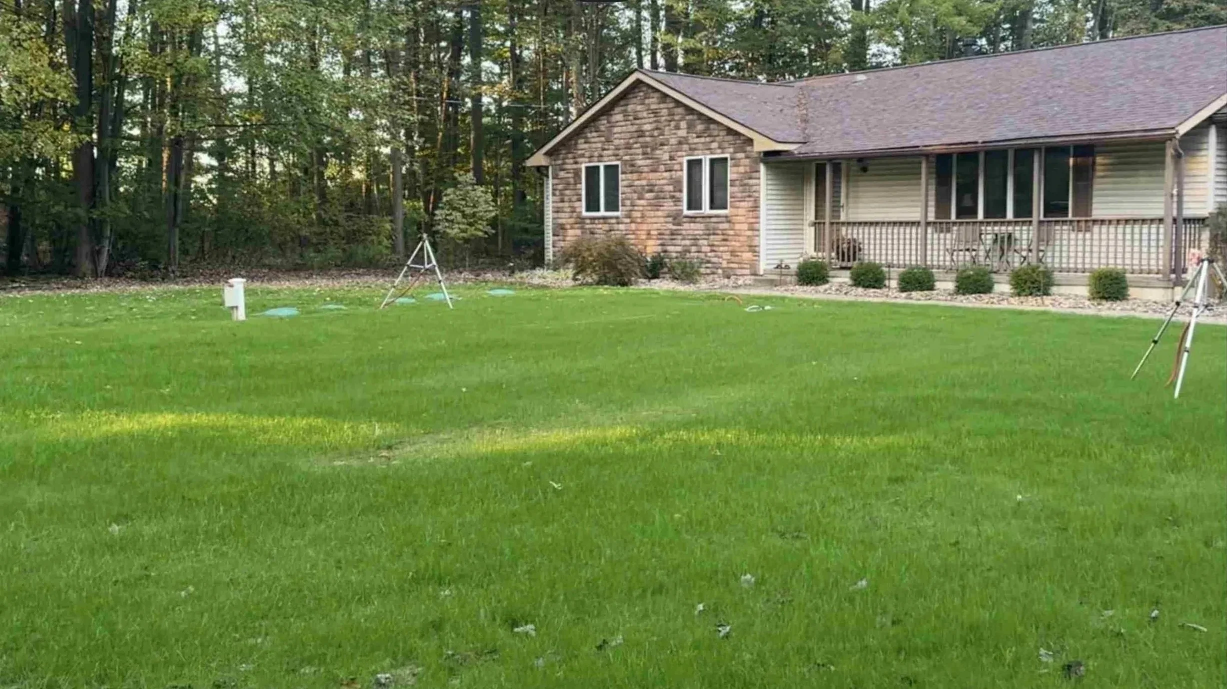 Front yard with a well-manicured green lawn, a house with brick and siding exterior, and a wooded treeline in the background.