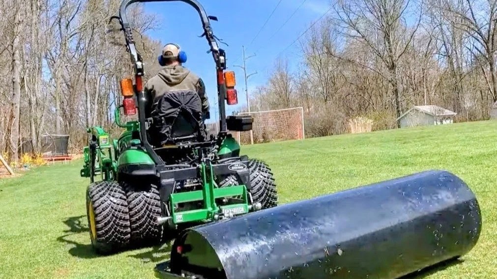 A person operating a green lawn roller on a grass field on a sunny day. The person is wearing headphones and a hoodie, and there is a soccer goal in the background.