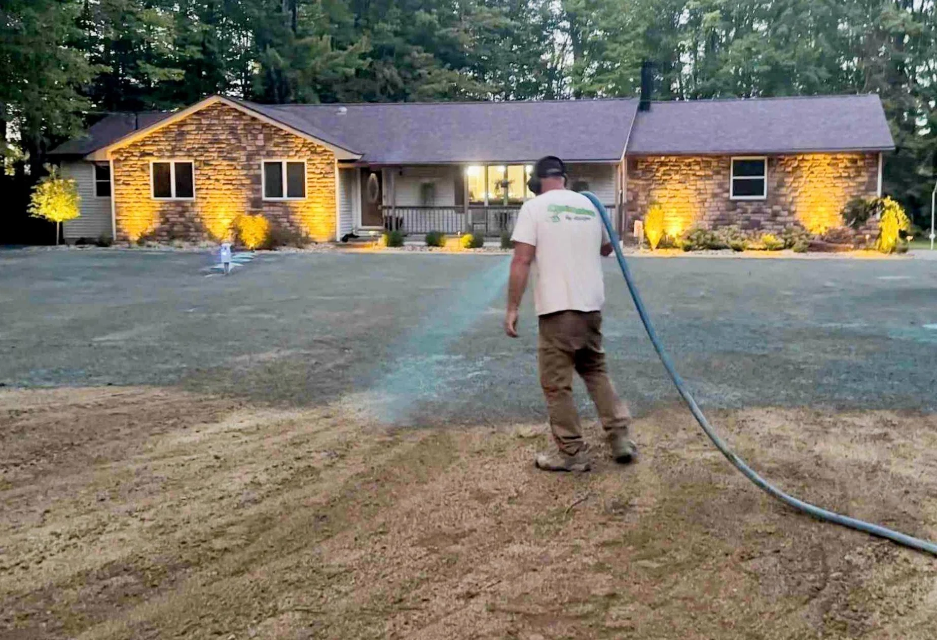 Person hydro seeding a front yard with a hose in front of a house with lit exterior lights and trees in the background.