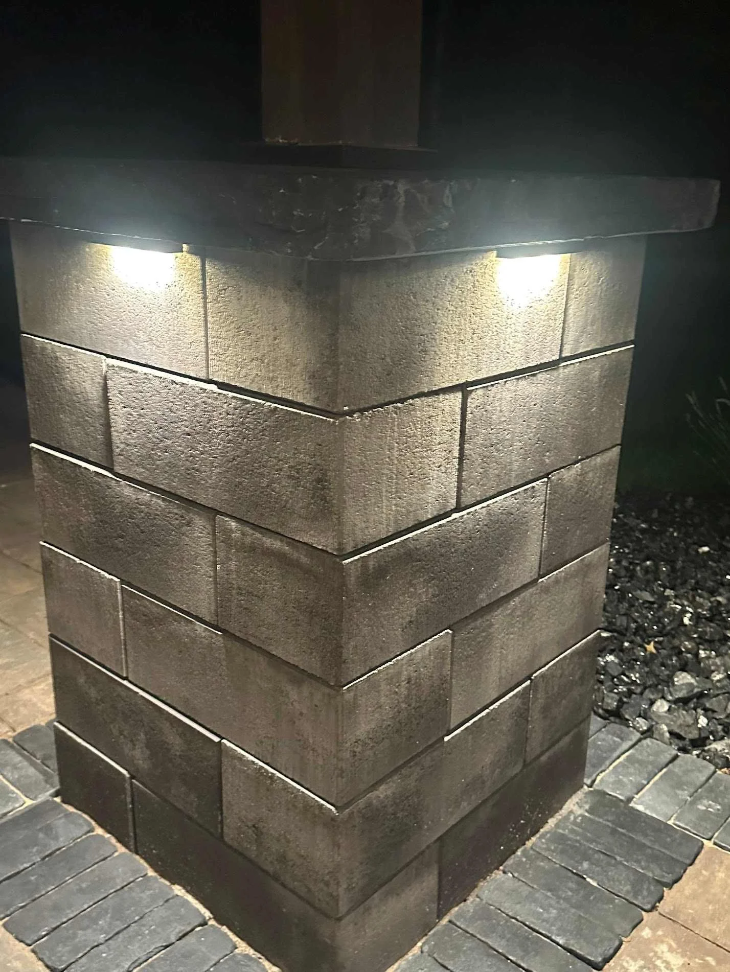 Close-up of a brick outdoor light post with illuminated lights, surrounded by dark ground and paving stones.