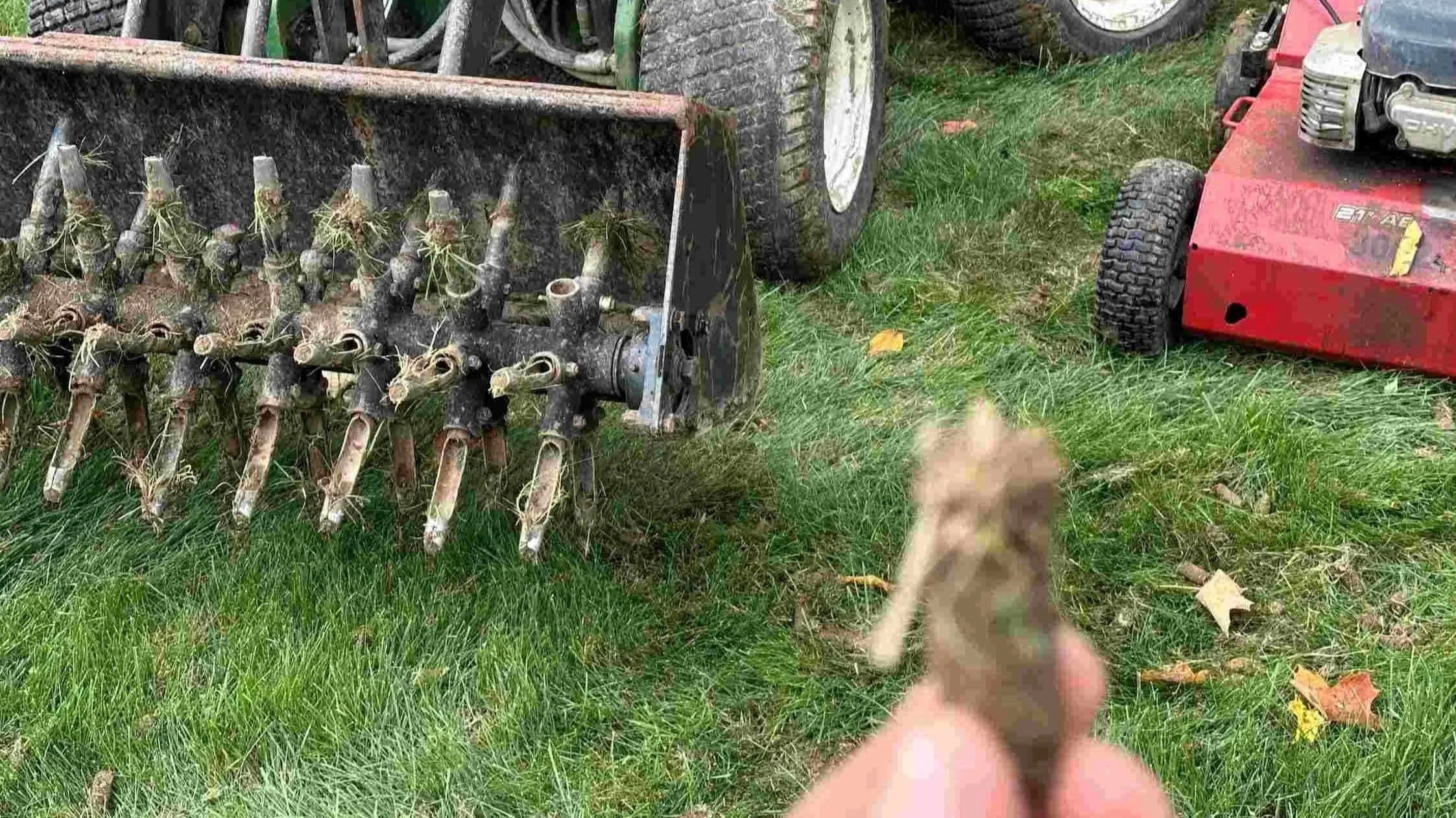 A person holding a small plant seedling in front of farm equipment and a red lawn mower on a grassy field.